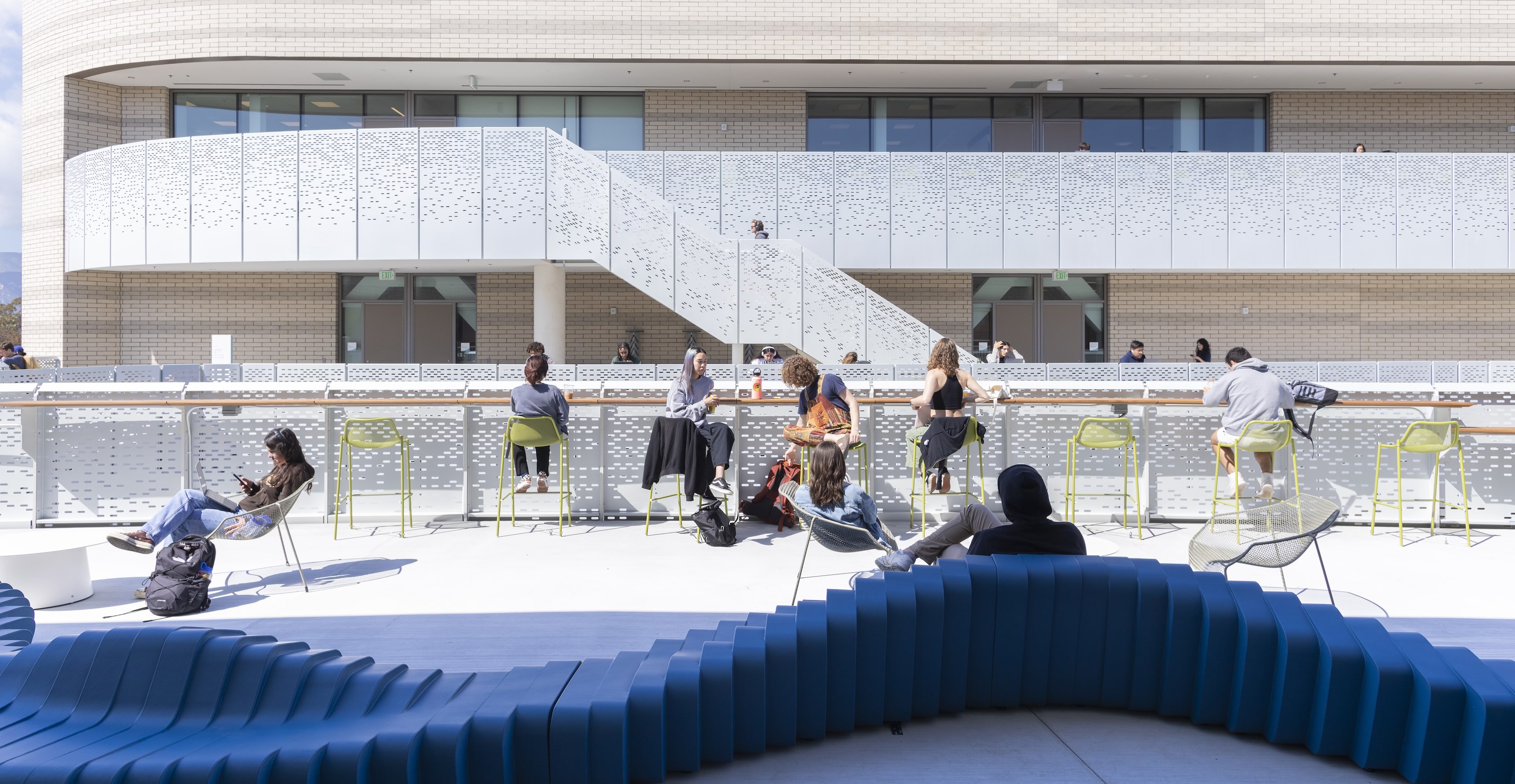 Students sitting in groups and talking on a long, curved blue bench in the sunny outdoor courtyard of the modern Interactive Learning Pavilion.